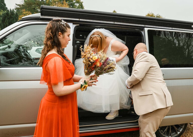 A bride getting out of her wedding vehicle with her dad helping and the bridesmaid dressed in bright orange holding the dried flowers on a wedding day at the Garden Barn, Little Bradley captured by Hayley Denston Photography