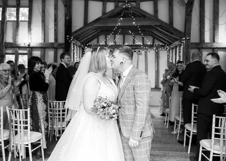 A black and white photo of a bride and groom kissing after they have walked down the aisle on a wedding day at the Garden Barn, Little Bradley captured by Hayley Denston Photography