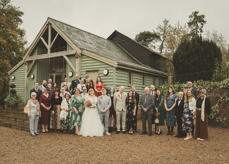 A large group photo of all the guests stood outside the front of a pale green barn on a wedding day at the Garden Barn, Little Bradley captured by Hayley Denston Photography