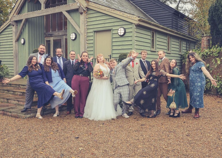 A fun group shot with everyone pulling silly faces in front of a pale green barn on a wedding day at the Garden Barn, Little Bradley captured by Hayley Denston Photography