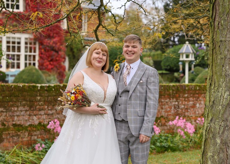 A bride and groom stood under a tree in autumnal gardens with beautiful foliage on their wedding day at the Garden Barn, Little Bradley captured by Hayley Denston Photography