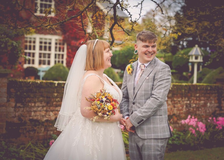 A bride and groom laughing under a tree in autumnal gardens with beautiful foliage on their wedding day at the Garden Barn, Little Bradley captured by Hayley Denston Photography