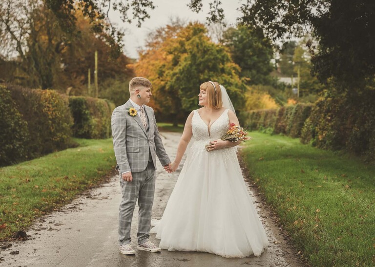A bride and groom stood in a road with autumnal trees in yellow and orange lining the sides of the road looking at each other on their wedding day at the Garden Barn, Little Bradley captured by Hayley Denston Photography