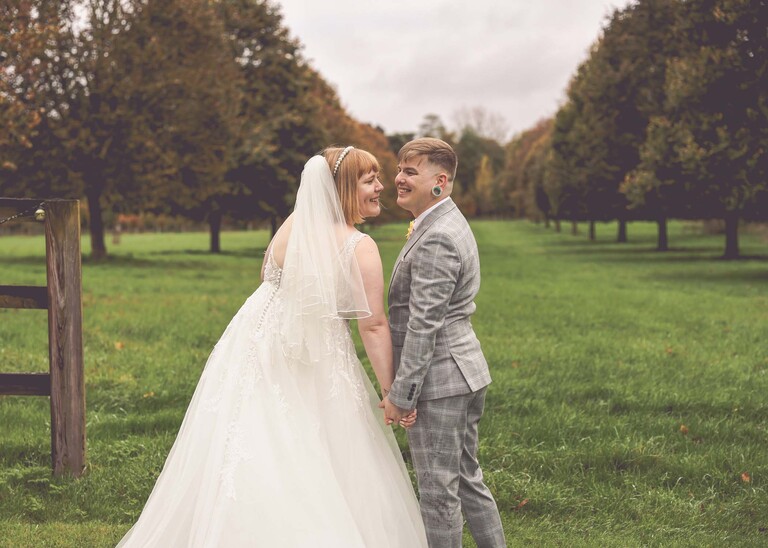 A bride and groom stood with their backs to the camera on a grass lawn looking back towards the camera laughing in front of a tree lined avenue on their wedding day at the Garden Barn, Little Bradley captured by Hayley Denston Photography
