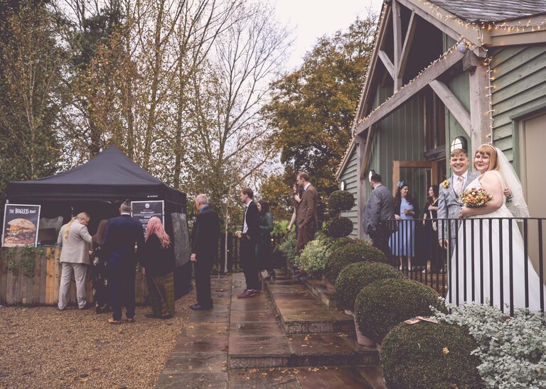 A bride and groom looking at the camera whilst guests line up for food from a food truck on a wedding day at the Garden Barn, Little Bradley captured by Hayley Denston Photography