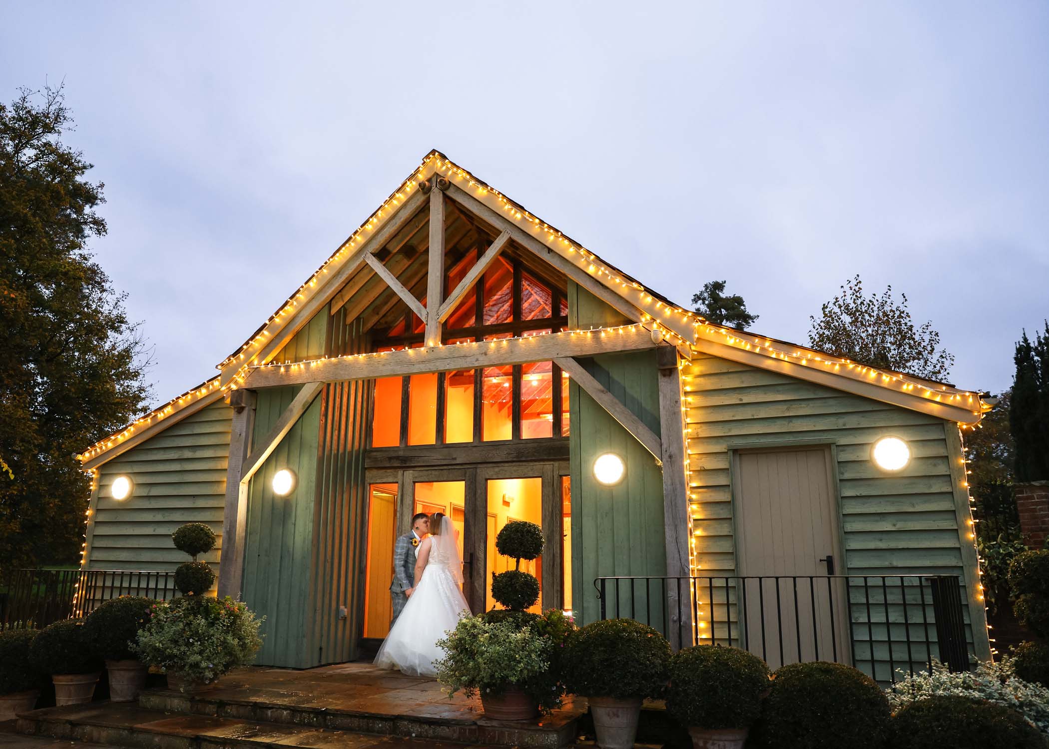 A bride and groom kissing in front of a barn lit with festoon lighting at dusk on their wedding day at the Garden Barn, Little Bradley captured by Hayley Denston Photography