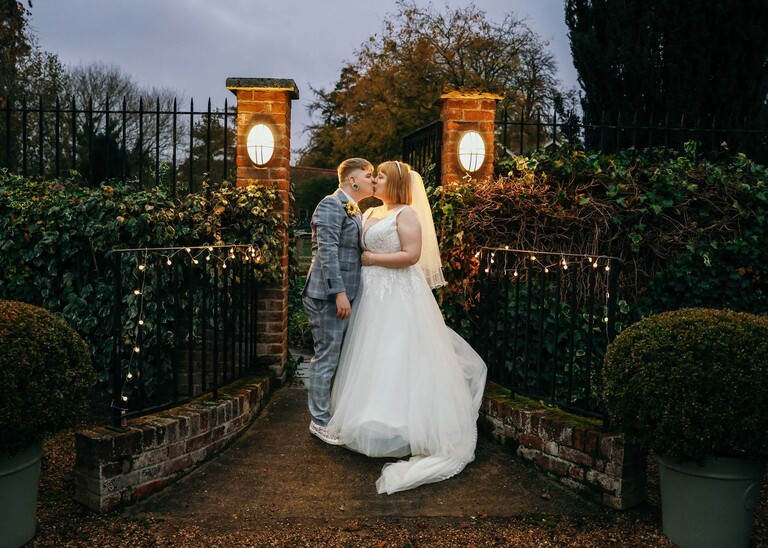 A bride and groom kissing at dusk in front of some wrought iron gates and lights on their wedding day at the Garden Barn, Little Bradley captured by Hayley Denston Photography