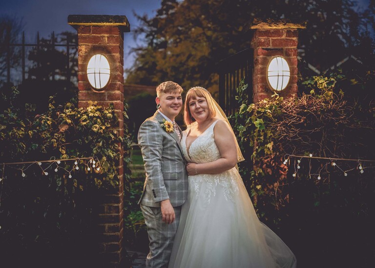 A bride and groom stood ina gateway surrounded by fairy lights and two main oval lights at dusck on their wedding day at the Garden Barn, Little Bradley captured by Hayley Denston Photography