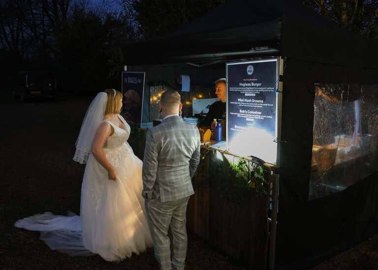 A bride and groom getting ready for their food and talking to the caterer in a food tent in the dark on their wedding day at the Garden Barn, Little Bradley captured by Hayley Denston Photography