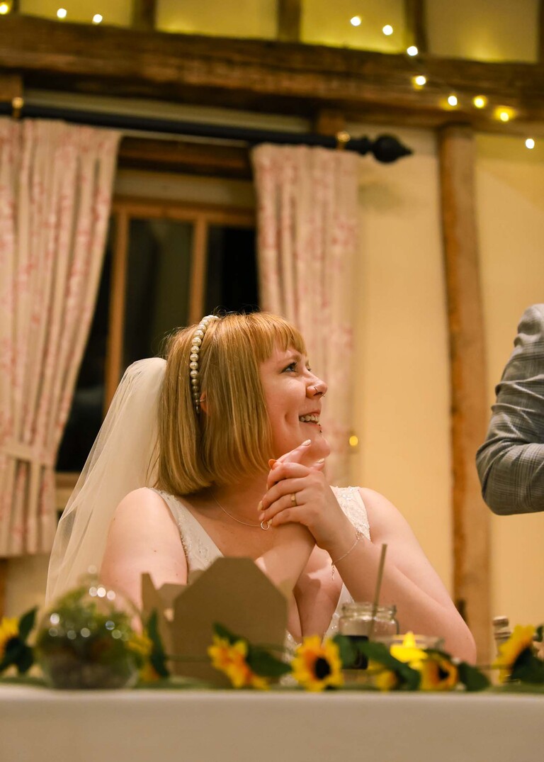 A bride looking up at her groom smiling during speeches on a wedding day at the Garden Barn, Little Bradley captured by Hayley Denston Photography