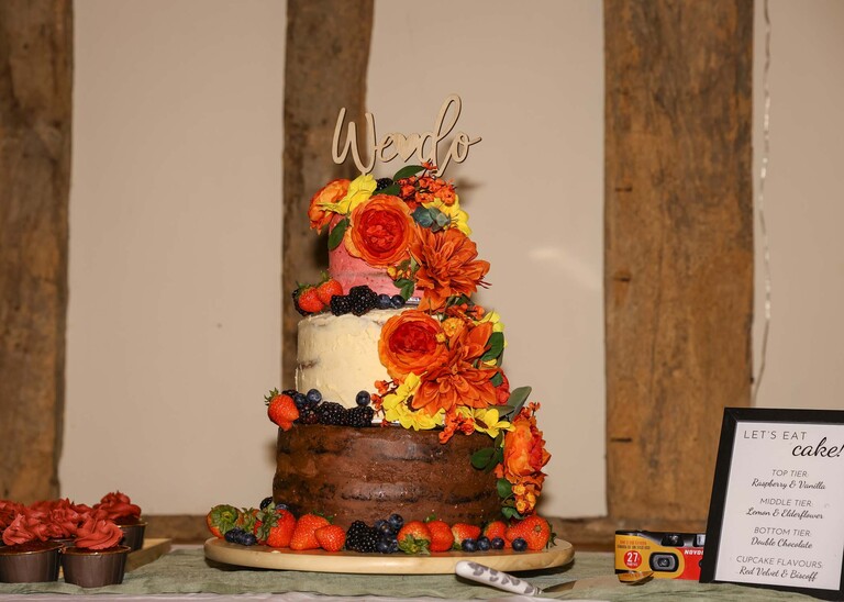 A wedding cake with a pink top layer, cream middle layer and chocolate bottom layer covered with bright flowers and berries on a wedding day at the Garden Barn, Little Bradley captured by Hayley Denston Photography