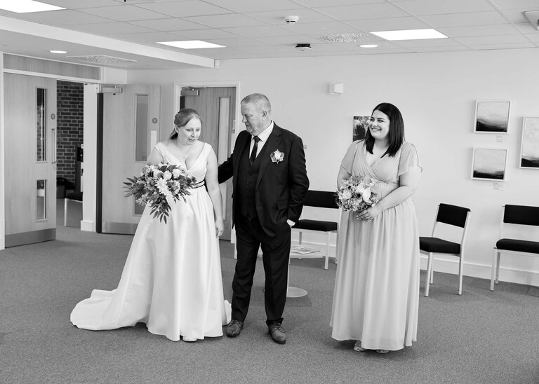 A black and white photo of a bride, her dad and bridesmaid waiting to come in to the wedding ceremony photographed by Suffolk Wedding Photographer Hayley Denston Photography