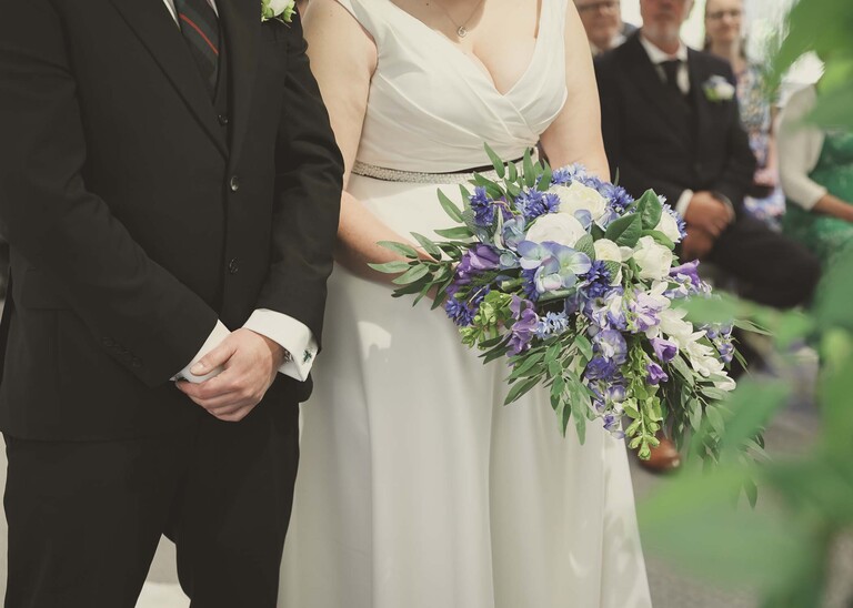 A close up of the groom and brides hands holding her bouquet during their wedding ceremony at Cambridge Registry Office photographed by Suffolk Wedding Photographer Hayley Denston Photography