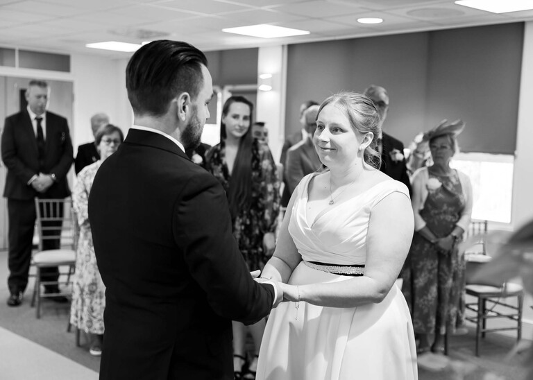 A black and white photo of a bride and groom looking at each other holding hands during their wedding ceremony at Cambridge Registry Office photographed by Suffolk Wedding Photographer Hayley Denston Photography