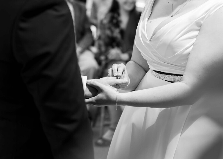 A black and white photo of the bride putting the grooms wedding ring on his finger during their wedding ceremony at Cambridge Registry Office photographed by Suffolk Wedding Photographer Hayley Denston Photography