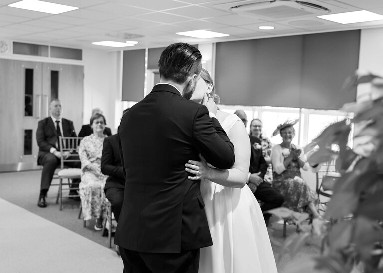 A black and white photo of a bride and groom sharing their first kiss during their wedding ceremony at Cambridge Registry Office photographed by Suffolk Wedding Photographer Hayley Denston Photography