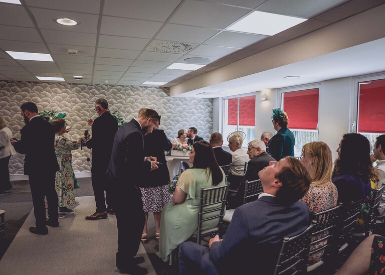 A bride and groom kissing after signing the register surrounded by guests taking photos during their wedding ceremony at Cambridge Registry Office photographed by Suffolk Wedding Photographer Hayley Denston Photography