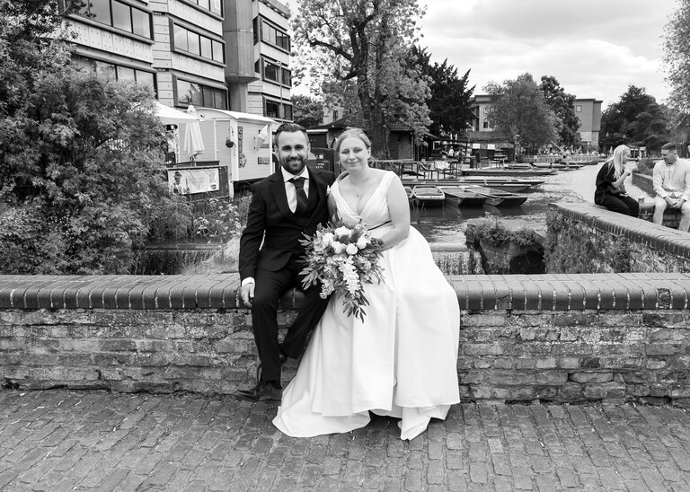 A black and white photo of a bride and groom sat on a wall at Cambridge Mill Pond on their wedding day photographed by Suffolk Wedding Photographer Hayley Denston Photography