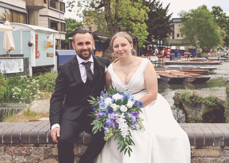 A photo of a bride and groom sat on a wall at Cambridge Mill Pond on their wedding day photographed by Suffolk Wedding Photographer Hayley Denston Photography