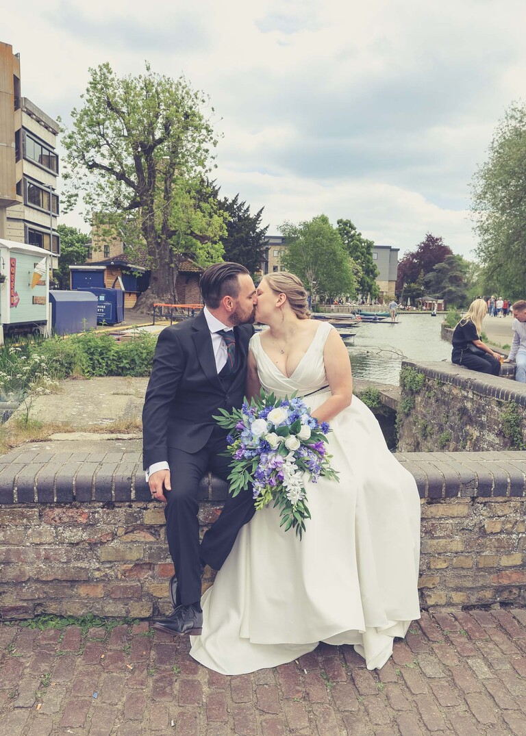 A photo of a bride and groom kissing sat on a wall at Cambridge Mill Pond on their wedding day photographed by Suffolk Wedding Photographer Hayley Denston Photography