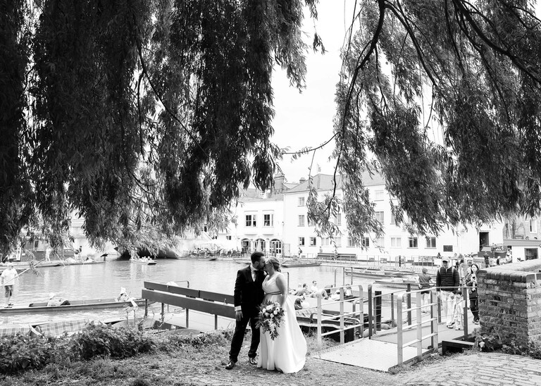 A black and white photo of a bride and groom kissing with the punts behind them through willow trees at Cambridge Mill Pond on their wedding day photographed by Suffolk Wedding Photographer Hayley Denston Photography