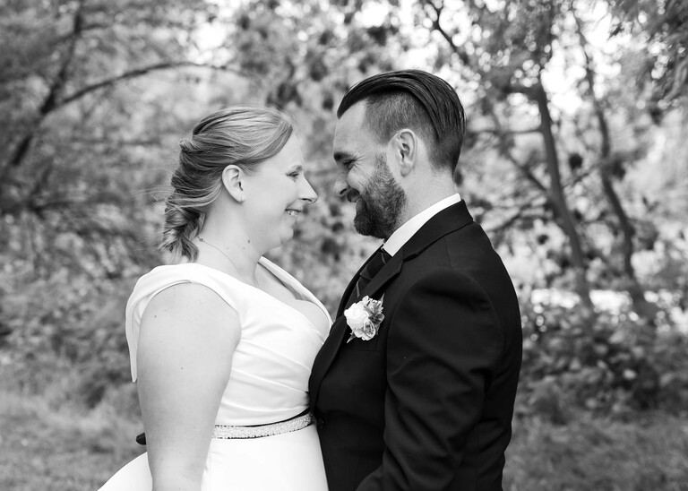 A black and white photo of a bride and groom looking at each other smiling with trees behind them in Meadows down at Cambridge Mill Pond on their wedding day photographed by Suffolk Wedding Photographer Hayley Denston Photography