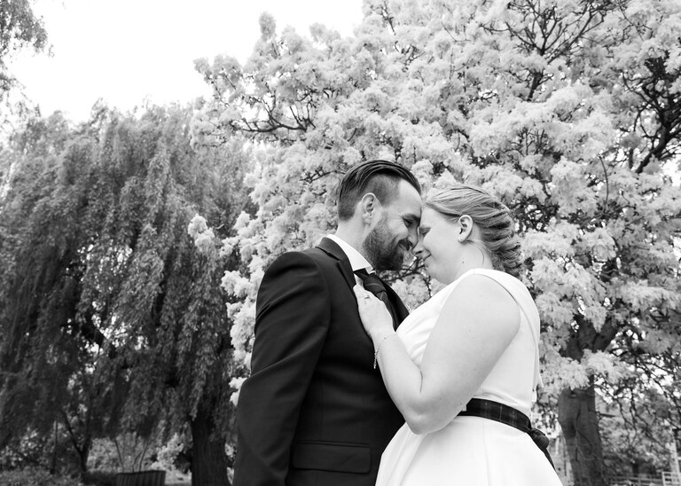 A black and white photo of a bride and groom with their heads together snuggling and trees behind in Meadows down at Cambridge Mill Pond on their wedding day photographed by Suffolk Wedding Photographer Hayley Denston Photography