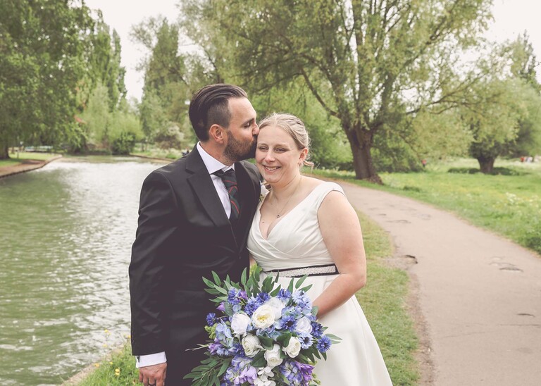 A groom kissing his bride on the head with the river cam behind them at Cambridge Mill Pond on their wedding day photographed by Suffolk Wedding Photographer Hayley Denston Photography