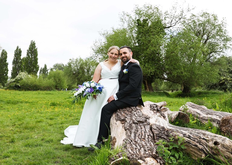 A bride and groom sat on a fallen tree trunk in Meadows down at Cambridge Mill Pond on their wedding day photographed by Suffolk Wedding Photographer Hayley Denston Photography