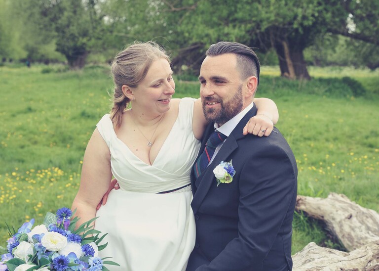 A bride sat on her grooms lap on a fallen tree in Meadows down at Cambridge Mill Pond on their wedding day photographed by Suffolk Wedding Photographer Hayley Denston Photography