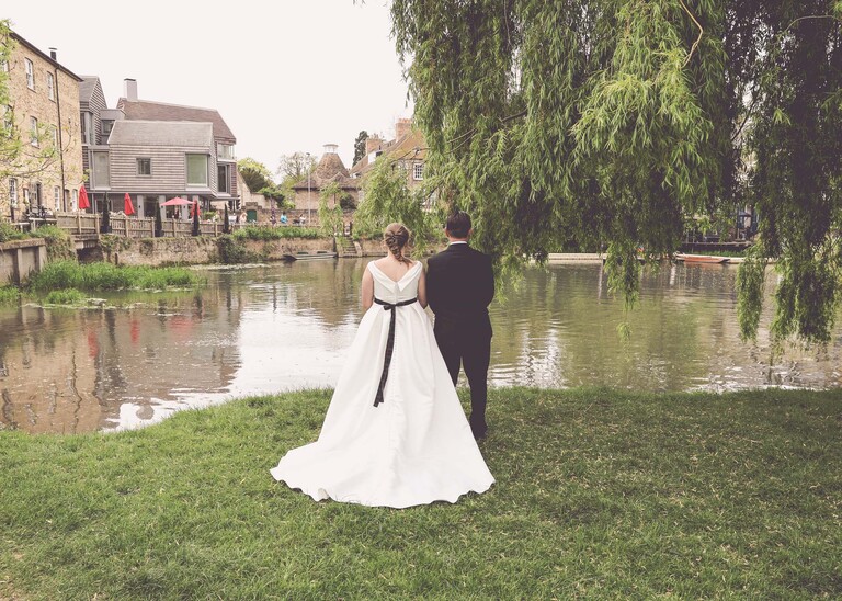 A bride and groom from behind with her dress laid out and the cambridge mill pond in front of them in on their wedding day photographed by Suffolk Wedding Photographer Hayley Denston Photography