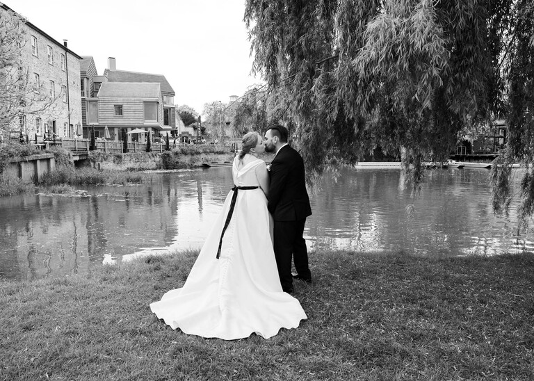 A black and white photo of a bride and groom kissing from behind with her dress laid out and the cambridge mill pond in front of them in on their wedding day photographed by Suffolk Wedding Photographer Hayley Denston Photography