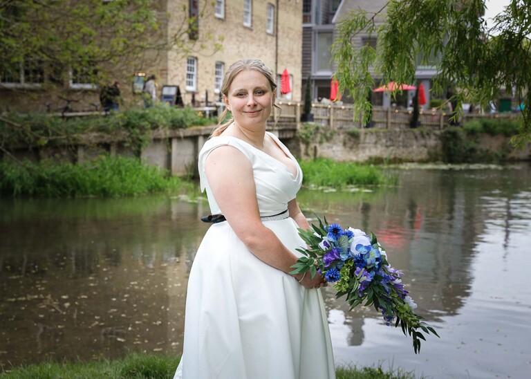 A bride smiling at the camera at cambridge mill pond in front of them in on their wedding day photographed by Suffolk Wedding Photographer Hayley Denston Photography
