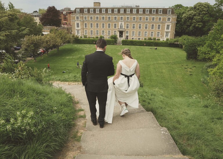 A bride and groom walking down Castle Hill in Cambridge from behind on their wedding day with the shire hall in front of them photographed by Suffolk Wedding Photographer Hayley Denston Photography