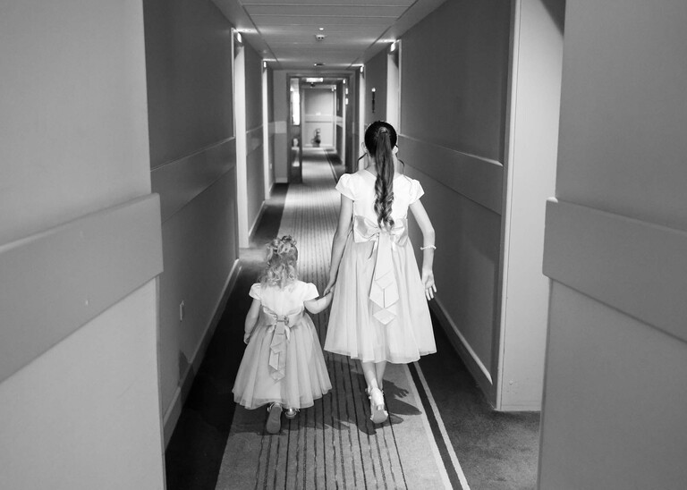 A black and white photo of two flowergirls walking away hand in hand down a hotel corridor on a wedding day captured by suffolk wedding Photographer Hayley Denston Photography