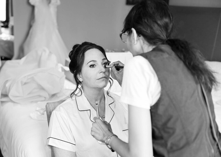 A black and white photo of a bride having her eyeshadow done by a make up artist on a wedding day captured by suffolk wedding Photographer Hayley Denston Photography