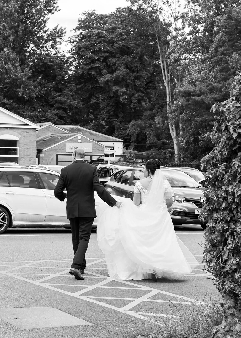 A black and white photo of the bride and her dad walking away to go get into her wedding vehicle on a wedding day captured by suffolk wedding Photographer Hayley Denston Photography