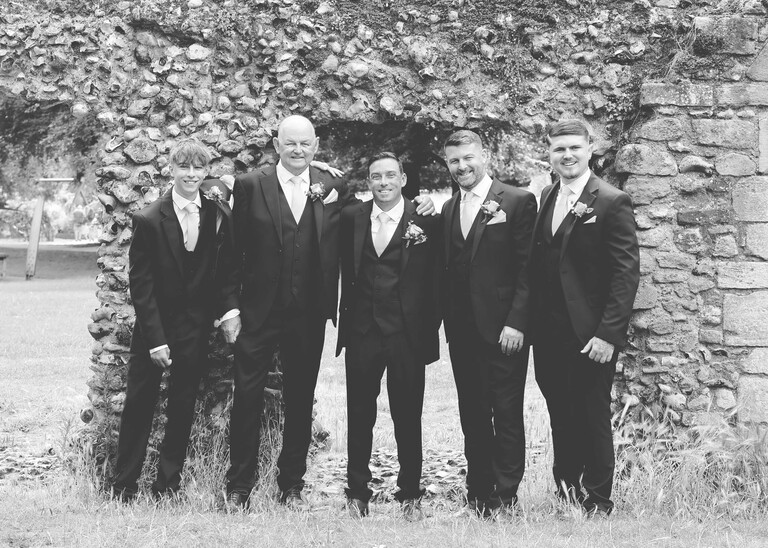 A black and white photo of the groom and his groomsmen stood in front of the abbey garden ruins in bury st edmunds on a wedding day captured by suffolk wedding Photographer Hayley Denston Photography