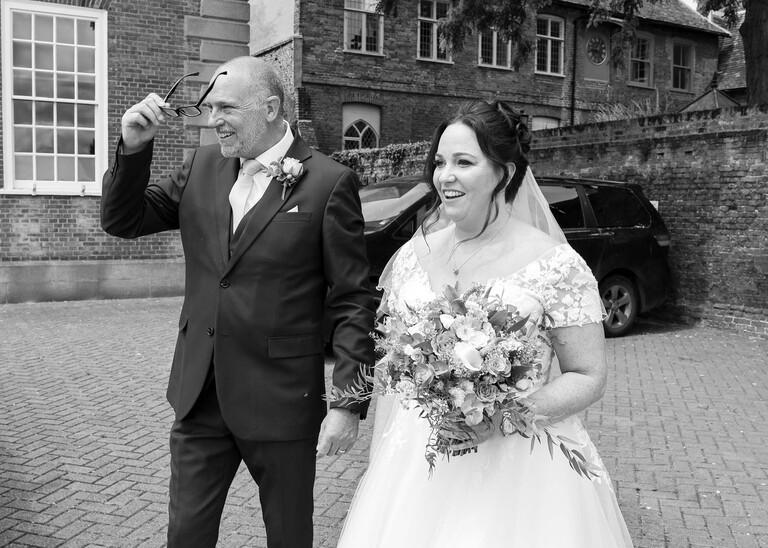 A black and white photo of a bride and her dad watching the bridal party arriving on a wedding day captured by suffolk wedding Photographer Hayley Denston Photography