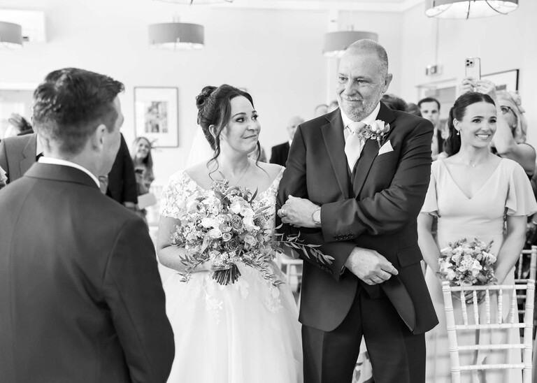 A black and white photo of a bride looking at her dad as they walk down the aisle at bury st edmunds registry office on a wedding day captured by suffolk wedding photographer Hayley Denston Photography