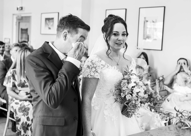 A black and white photo of a groom wiping away a tear with a hankie stood next to his bride during their wedding ceremony at bury st edmunds registry office on a wedding day captured by suffolk wedding photographer Hayley Denston Photography