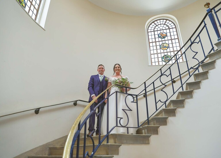 A bride and groom stood on the staircase with stained glass windows behind them after their wedding ceremony at bury st edmunds registry office on a wedding day captured by suffolk wedding photographer Hayley Denston Photography
