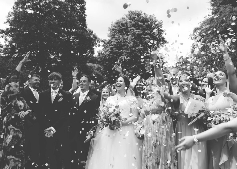 A black and white photo of a bride and groom stood amongst their guests with confetti being thrown on them in bury st edmunds abbey gardens on a wedding day captured by suffolk wedding photographer Hayley Denston Photography