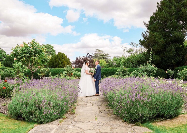 A bride and groom stood holding hand in hand in amongst lavender in bury st edmunds abbey gardens on a wedding day captured by suffolk wedding photographer Hayley Denston Photography