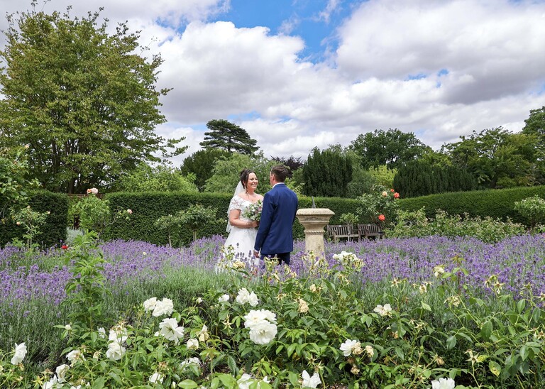 A bride stood looking at her groom surrounded by white roses and lavendar and blue cloudy skies in bury st edmunds abbey gardens on a wedding day captured by suffolk wedding photographer Hayley Denston Photography