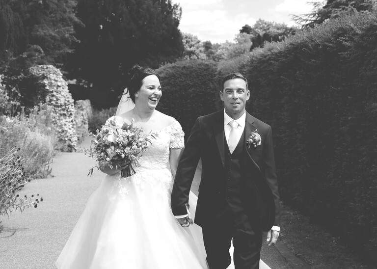 A black and white photo of a bride looking at her groom as they walk hand in hand through bury st edmunds abbey gardens on a wedding day captured by suffolk wedding photographer Hayley Denston Photography