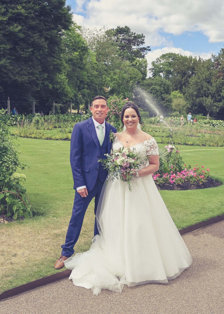 A bride and groom stood in front of flowerbeds with a water fountain going behind them in bury st edmunds abbey gardens on a wedding day captured by suffolk wedding photographer Hayley Denston Photography