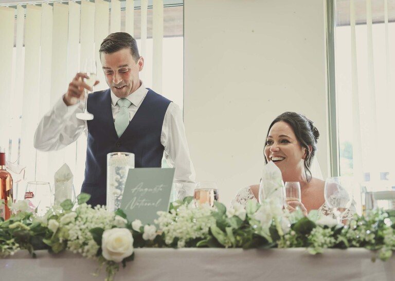 A groom raising a toast with the bride sat next to him during speeches at Mildenhall Social Club for a wedding reception captured by Suffolk Wedding Photographer Hayley Denston Photography