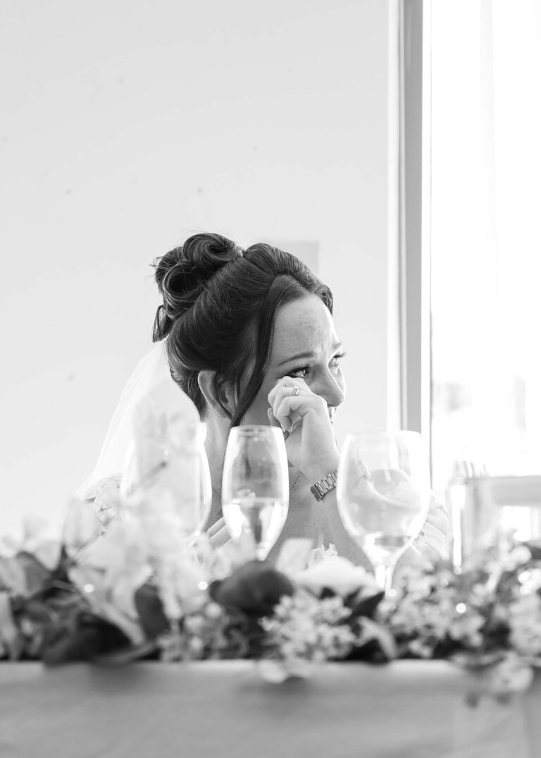 A black and white photo of a bride wiping away a tear during speeches at Mildenhall Social Club for a wedding reception captured by Suffolk Wedding Photographer Hayley Denston Photography