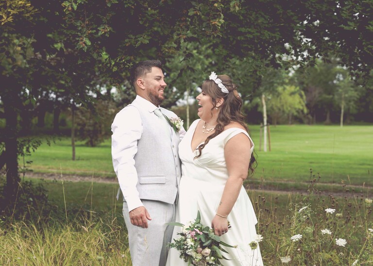 A bride and groom laughing at each other in long grass with trees behind them on their wedding day at Hungarian Hall Estate captured by Suffolk Wedding Photographer Hayley Denston Photography